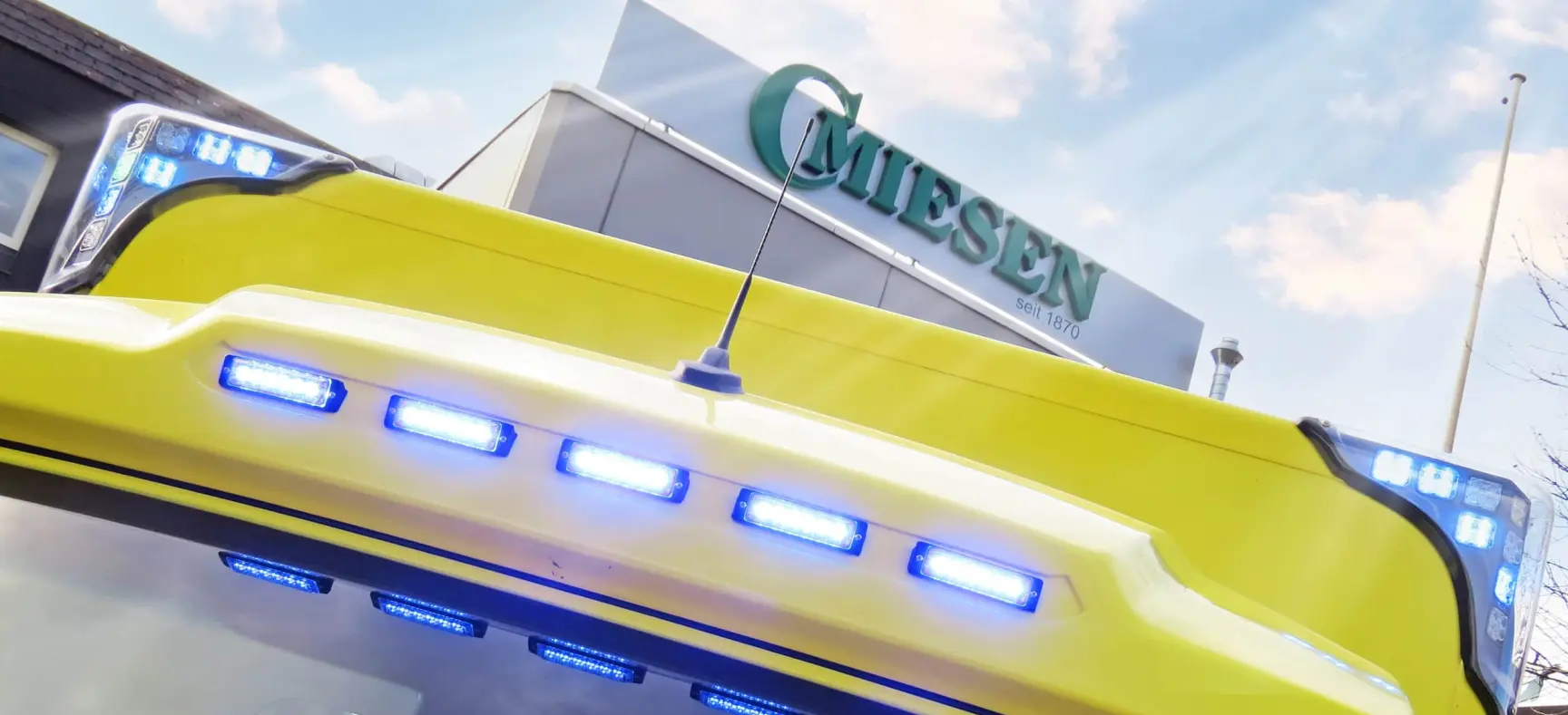 Frontansicht eines gelben Sprinter-ITW mit eingeschaltetem Blaulicht, vor Miesen-Firmengebäude.