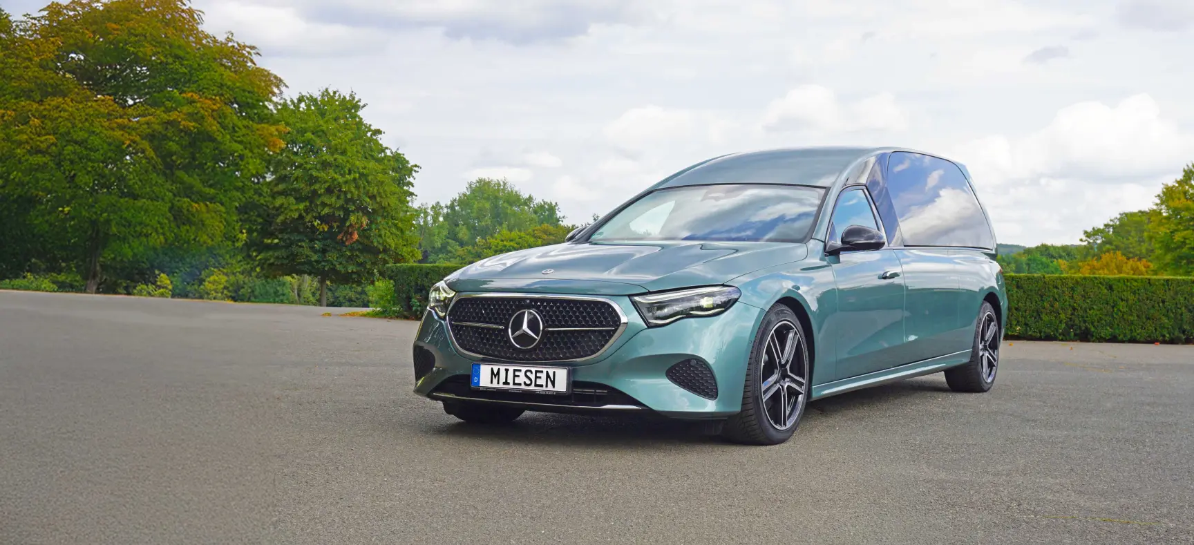 Grünes Fahrzeug mit Mercedes-Stern auf freier Fläche, umgeben von Bäumen und bewölktem Himmel.