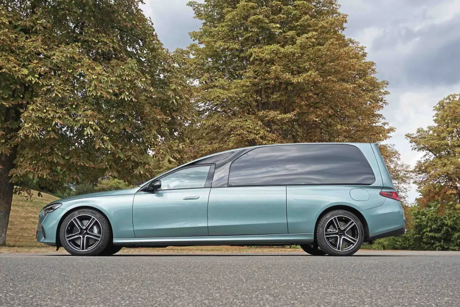 Seitenansicht eines türkisfarbenen Bestattungsfahrzeugs vor herbstlicher Baumkulisse.