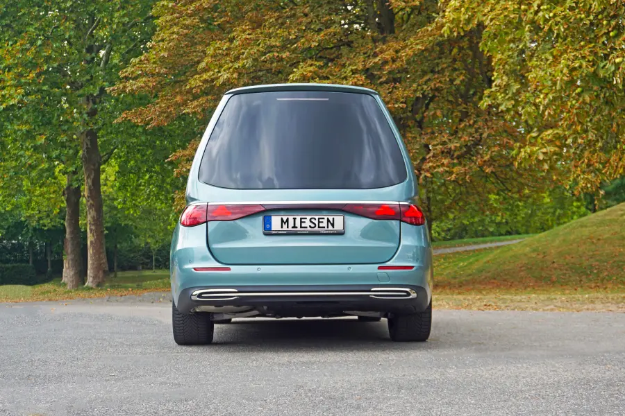 Rückansicht eines türkisfarbenen Fahrzeugs mit getönten Scheiben und dem Kennzeichen „Miesen“, geparkt in einer grünen Parklandschaft.