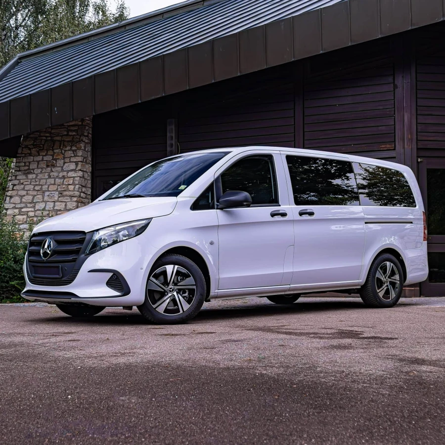 Weißer Bestattungswagen auf Basis eines Mercedes-Benz Vito, geparkt vor einem Gebäude mit dunkler Holzfassade und Natursteinmauerwerk; elegantes Design mit abgedunkelten Scheiben und stilvollen Leichtmetallfelgen.