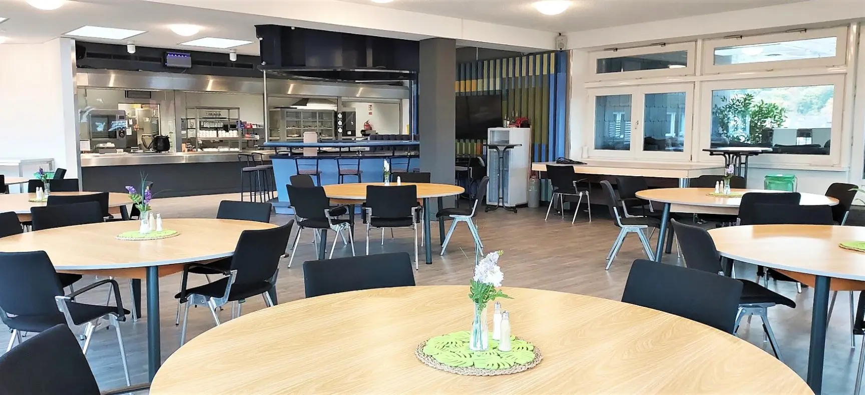 Moderne Kantine mit runden Holztischen, schwarzen Stühlen und offener Küchentheke im Hintergrund.