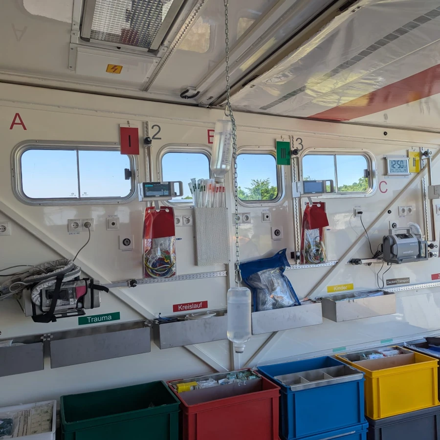 Innenansicht eines mobilen Behandlungsraums mit medizinischen Geräten, Hängeinfusion und farblich sortierten Materialboxen.