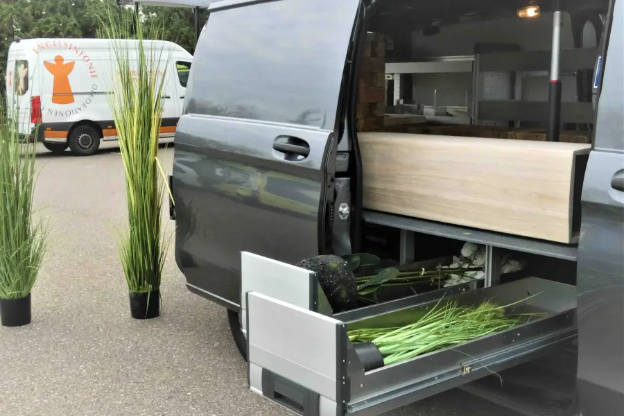 Seitenansicht eines Bestattungsfahrzeugs mit geöffnetem Auszugssystem und Dekorationselementen wie Gräsern und Pflanzen.