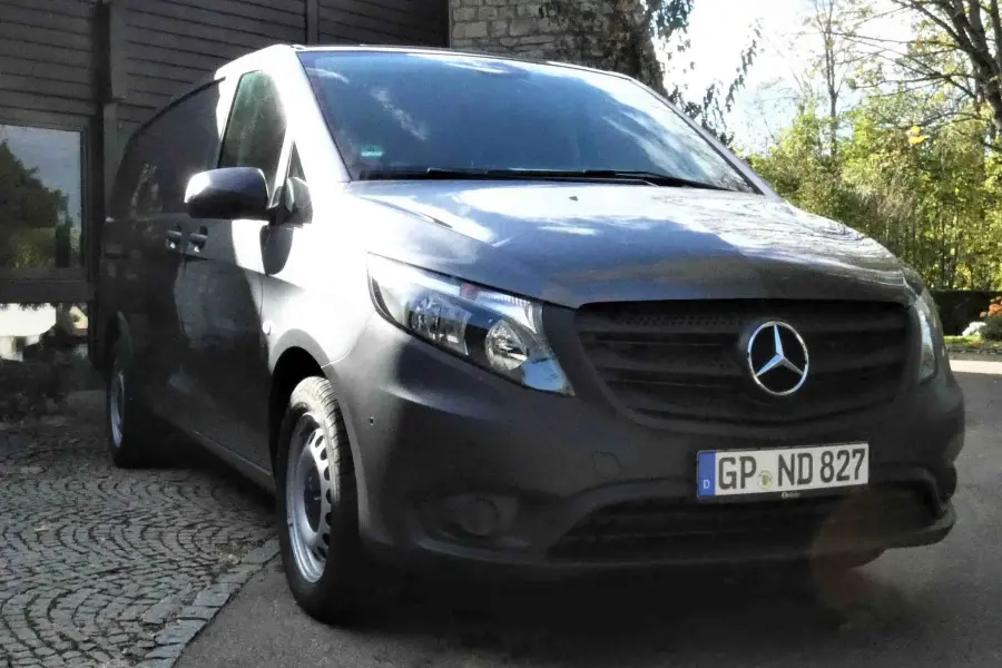 Frontansicht eines grauen Mercedes-Benz Transporters, geparkt vor einem Gebäude im Grünen.