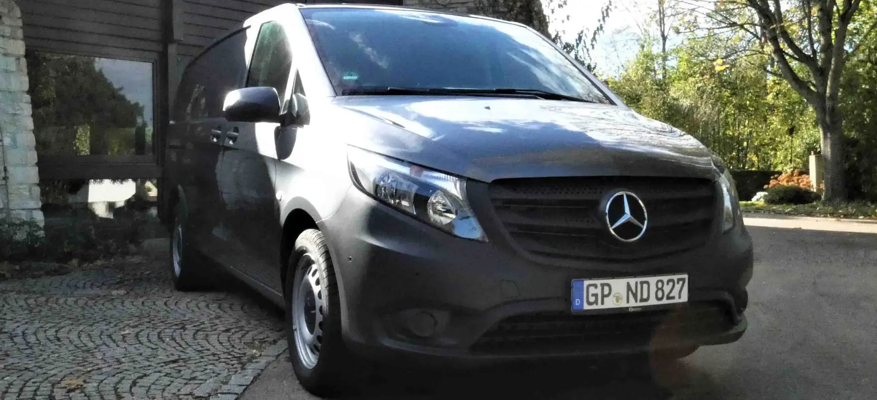 Frontansicht eines grauen Mercedes-Benz Transporters, geparkt vor einem Gebäude im Grünen.