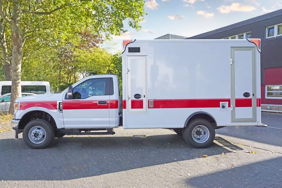 Seitenansicht eines weiß-roten Rettungswagens mit Kastenaufbau, geparkt unter Bäumen.
