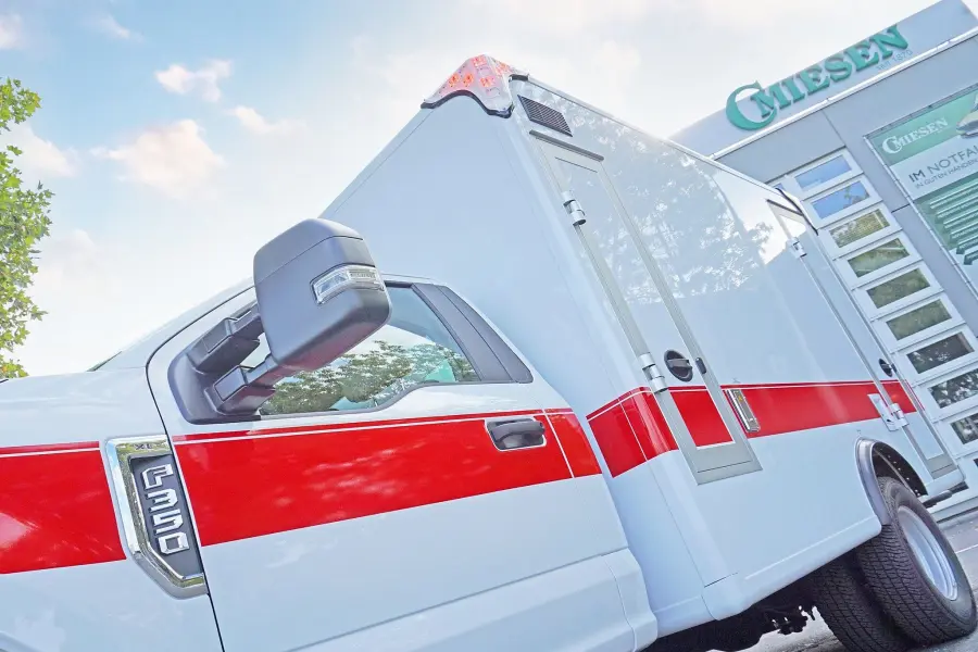 Seitenansicht eines weiß-roten Rettungswagens mit Kastenaufbau, geparkt unter Bäumen.