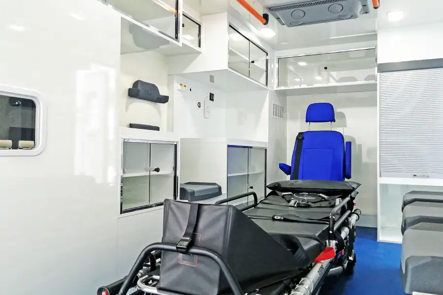 Seitenansicht eines weiß-roten Rettungswagens mit Kastenaufbau, geparkt unter Bäumen.