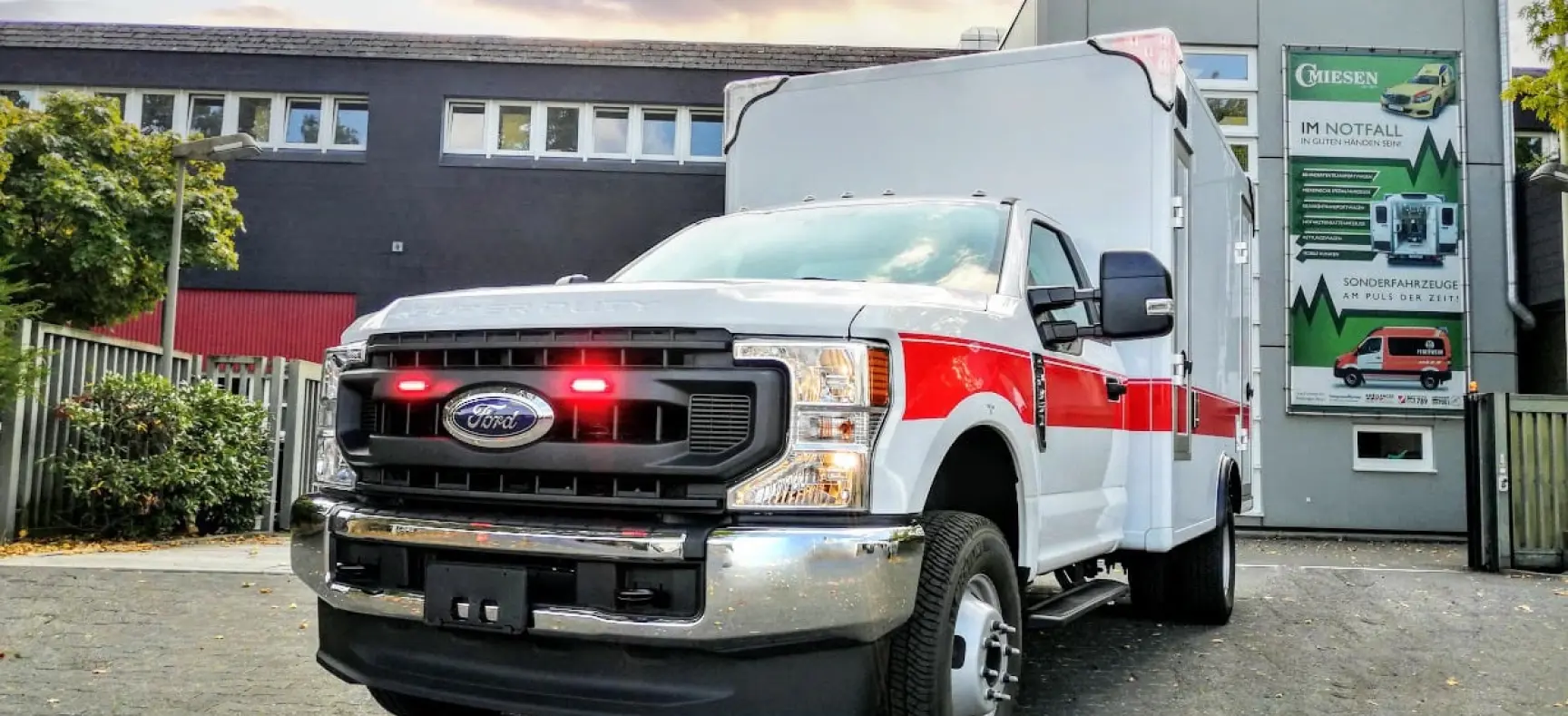 Frontansicht eines weißen Rettungswagens mit rotem Streifen und eingeschaltetem Blaulicht vor dem CMiesen-Gebäude.