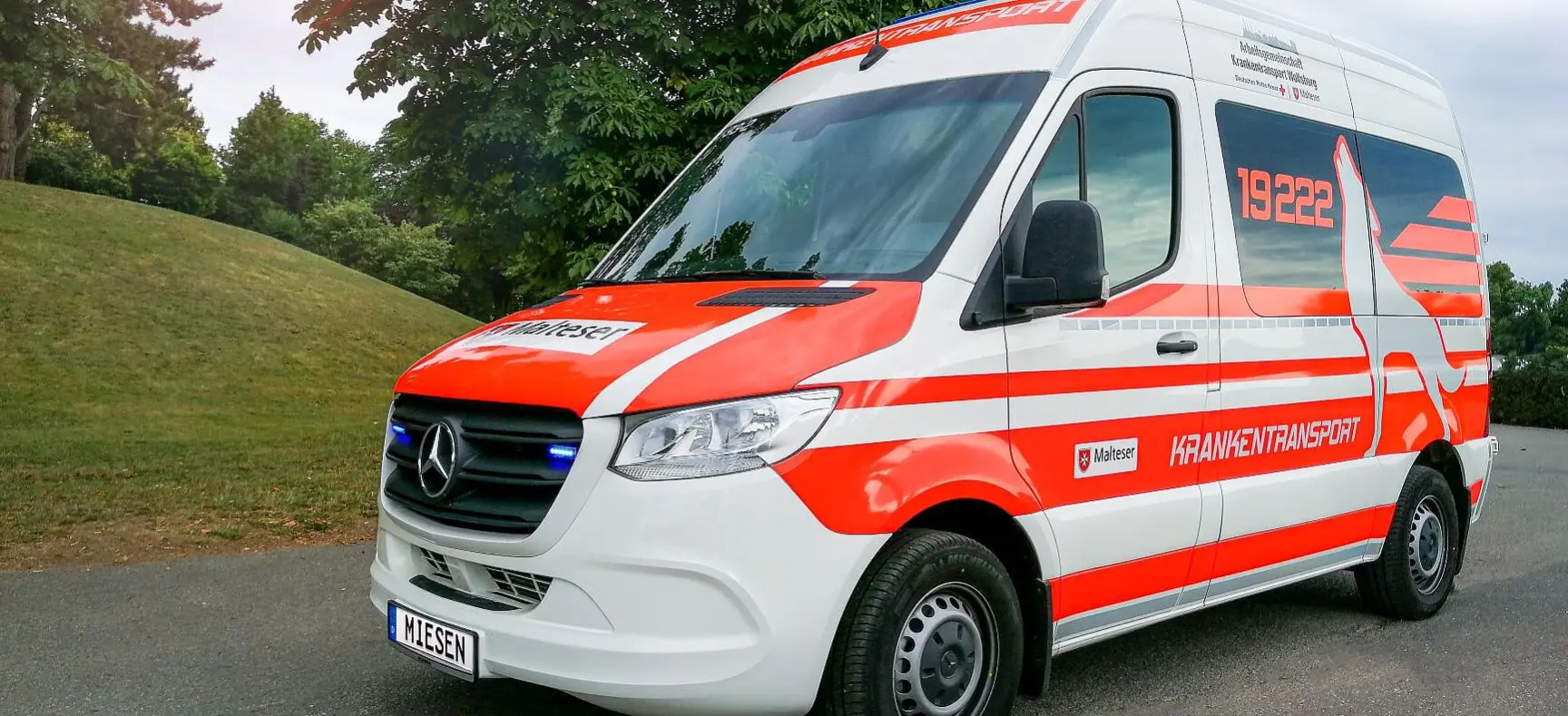 Krankentransportwagen der Malteser im Außenbereich, mit roter und weißer Beklebung und eingeschaltetem Blaulicht.