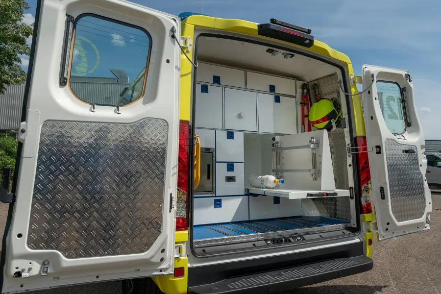 Geöffnete Hecktüren eines gelb-weißen Einsatzfahrzeugs mit übersichtlichem Stauraum, Schubladen und medizinischer Ausrüstung im Heckbereich.