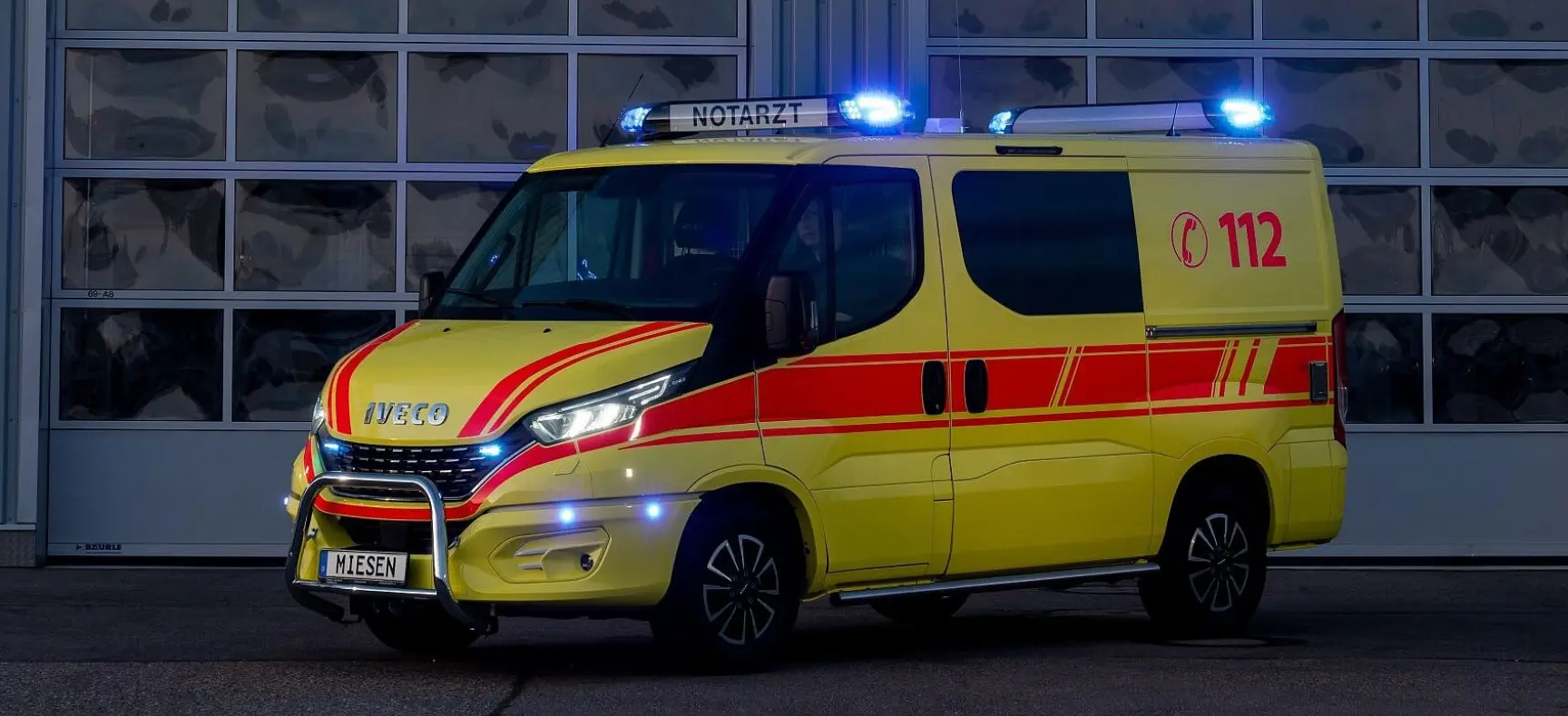 Gelber Notarztwagen mit roter Beklebung und eingeschaltetem Blaulicht, geparkt vor einer Fahrzeughalle bei Nacht.