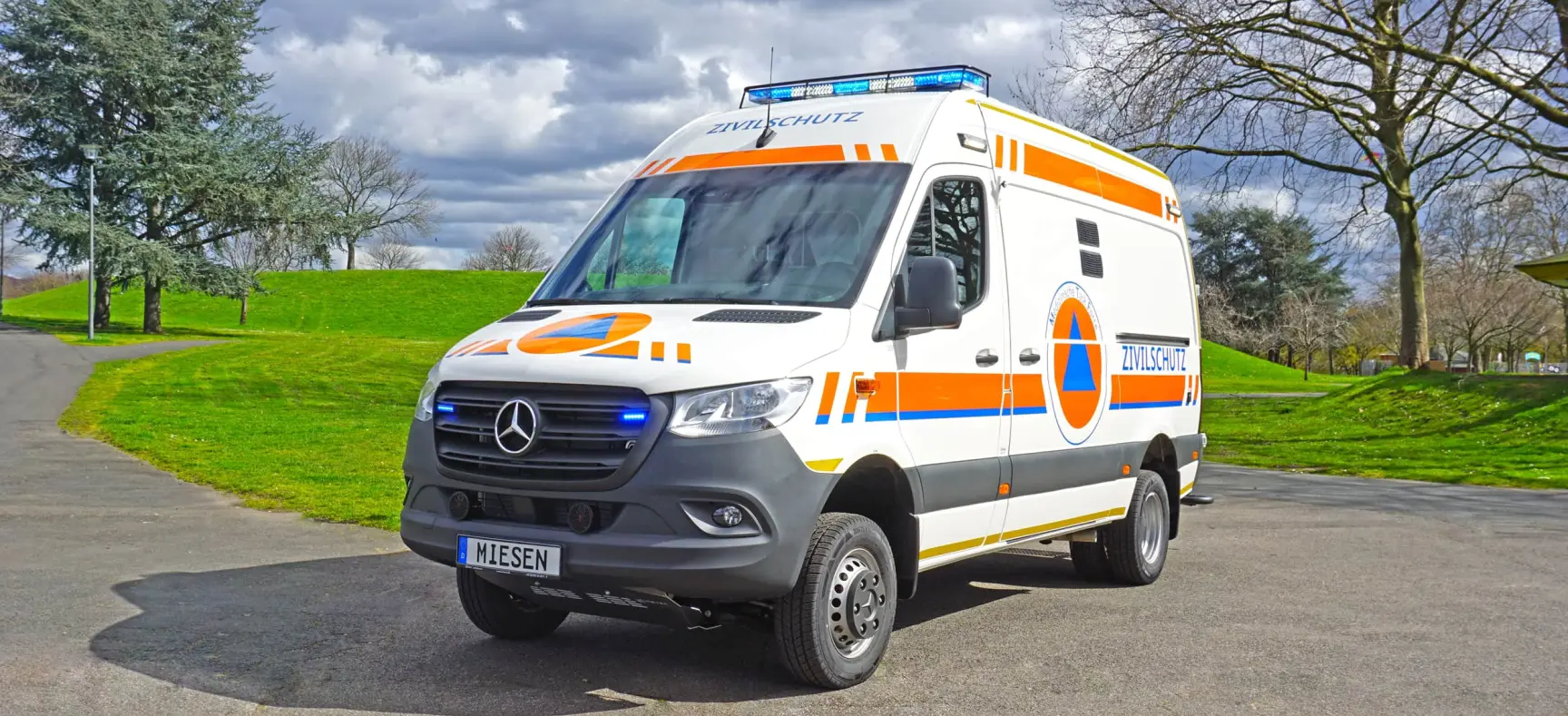 KTW des Zivilschutzes auf Mercedes-Basis mit orangener Beklebung, Blaulicht und Zivilschutz-Logo, geparkt auf Asphalt vor grüner Parklandschaft.