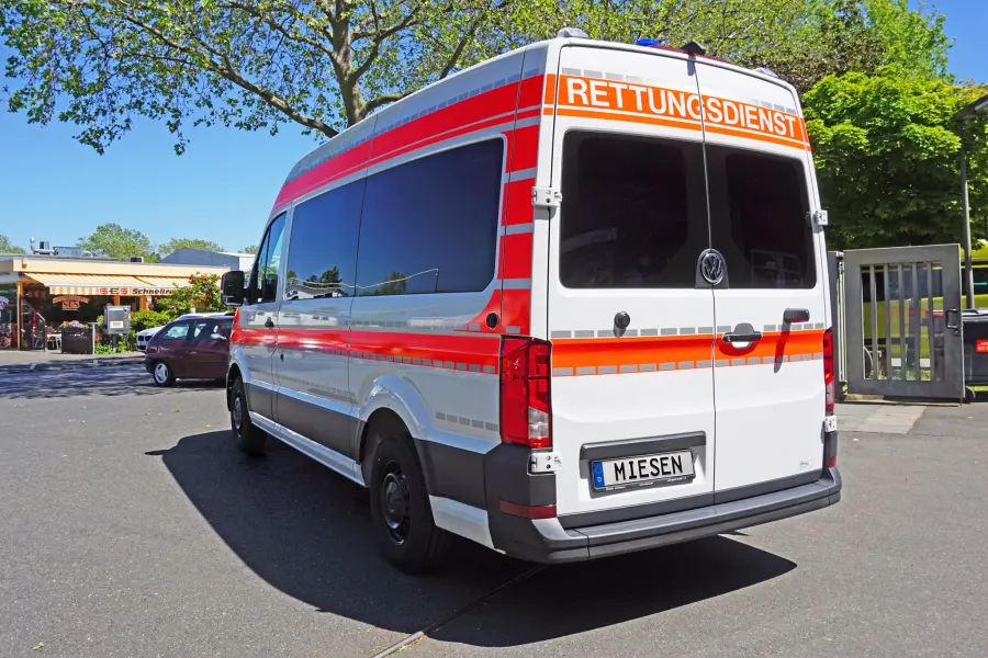 Heckansicht eines weißen Rettungswagens mit roter Beschriftung „Rettungsdienst“ und Miesen-Kennzeichen, geparkt auf einem sonnigen Parkplatz.