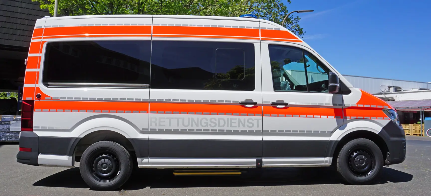 Rettungsdienstfahrzeug mit orange-grauer Beklebung, geparkt im Freien bei sonnigem Wetter.