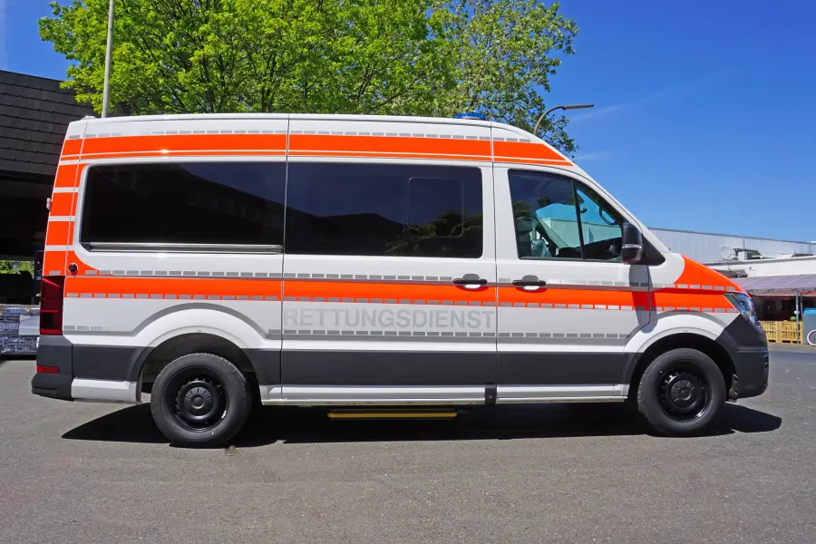 Rettungsdienstfahrzeug mit orange-grauer Beklebung, geparkt im Freien bei sonnigem Wetter.