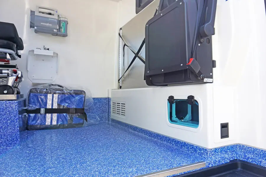 Innenraum eines Krankentransportwagens mit blauem Bodenbelag und zusammengeklapptem Sitzplatz.