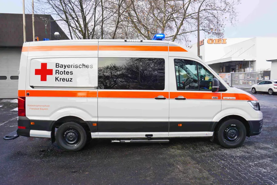 Seitenansicht eines Rettungswagens des Bayerischen Roten Kreuzes mit orangefarbener Warnbeklebung und Blaulicht.