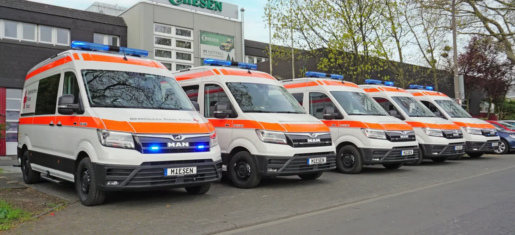 Mehrere KTW auf MAN TGE-Basis mit BRK-Beschriftung und Blaulicht, aufgereiht vor dem Firmengebäude von Miesen.