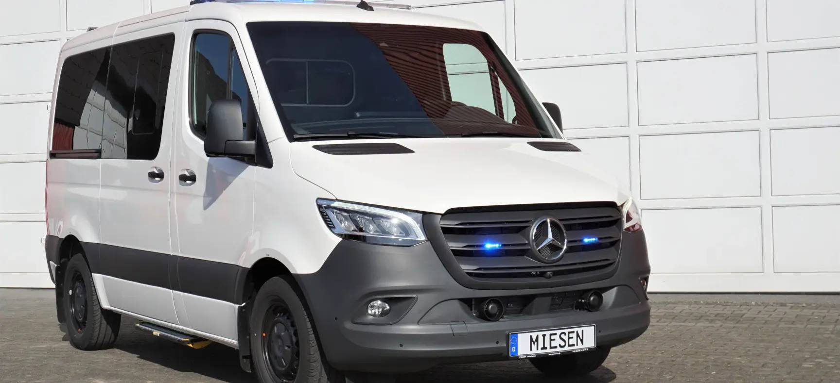 Weißer Mercedes-Benz Sprinter mit eingeschalteter blauer LED-Signalanlage vor einer Garagenwand.