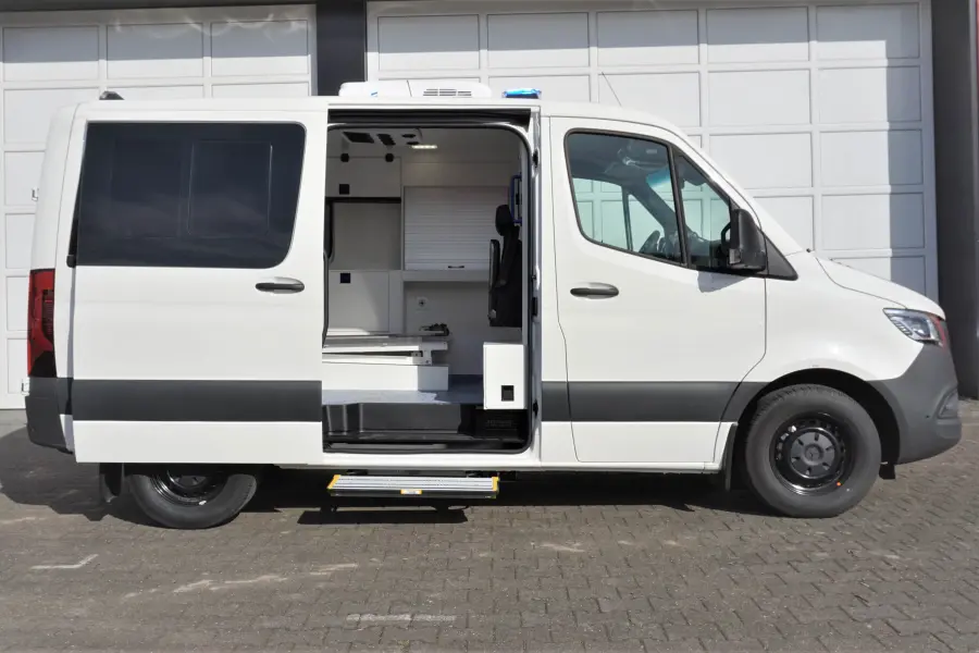 Weißer Krankentransportwagen mit geöffneter Seitentür und sichtbarem Innenraum vor einer Garage.