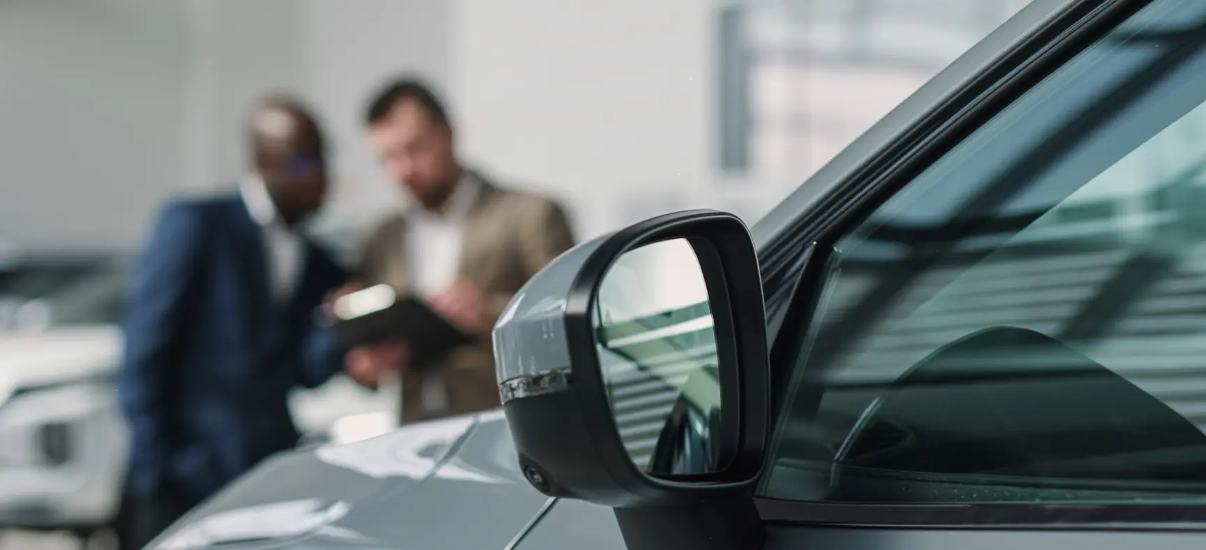 Seitenspiegel eines Autos im Vordergrund, unscharf im Hintergrund zwei Personen im Gespräch.