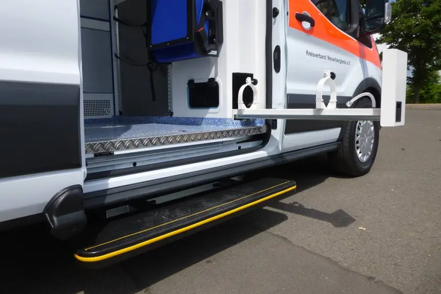 Seitliche Einstiegstufe eines Rettungswagens mit ausziehbarer Halterung und Blick ins teilweise geöffnete Fahrzeuginnere.