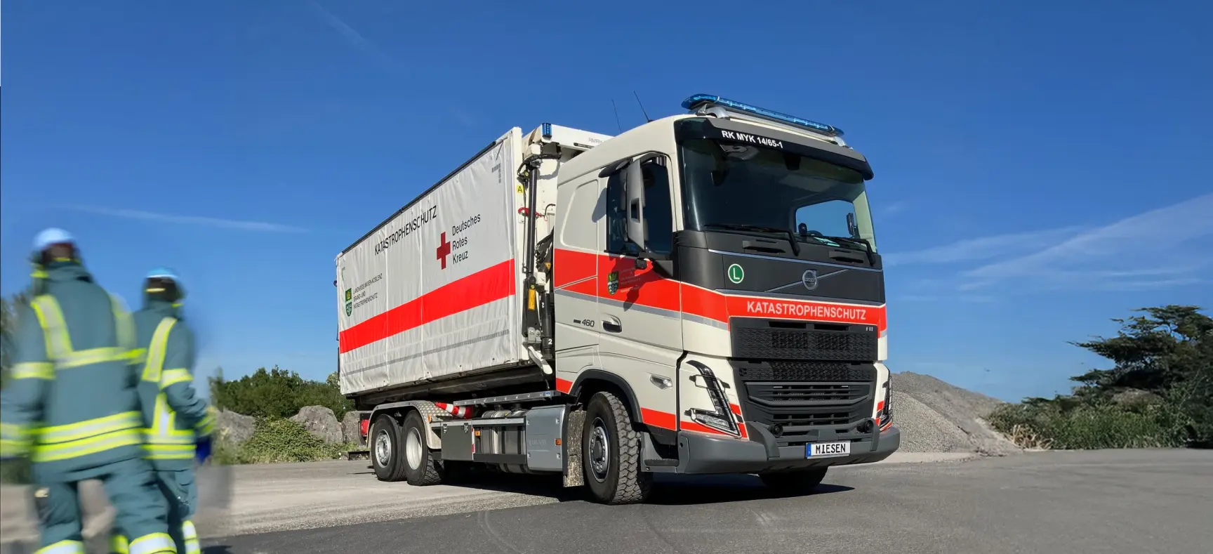 Lastwagen des Katastrophenschutzes mit Anhänger des Deutschen Roten Kreuzes auf einer Straße, zwei Einsatzkräfte im Vordergrund.