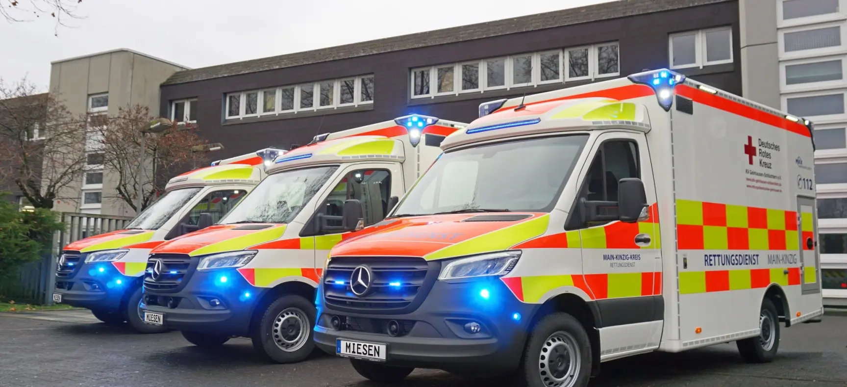 Drei Rettungswagen des Deutschen Roten Kreuzes mit eingeschaltetem Blaulicht vor einem modernen Gebäude.